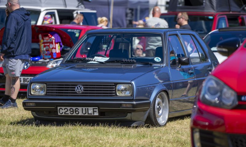 A blue VW Golf CL sits on display in the show and shine at Mighty Dub Fest on Alnwick Castle Pastures, with other cars peeking into the photo